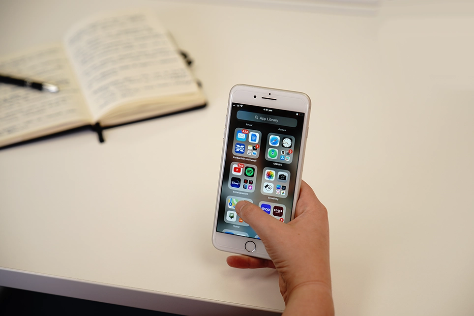 Hand holding an Iphone mobile phone with the app library open and with a notebook and pen in the background